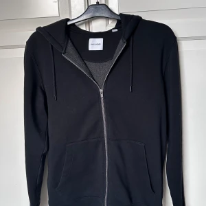 Svart hoodie från Jack & Jones - Snygg svart hoodie från Jack & Jones med dragkedja och justerbar huva. Perfekt för en avslappnad stil. Den har långa ärmar och praktiska fickor framtill. Fraktar gärna, finns och hämta i Bollebygd annars.                                              Hör av er vid frågor!