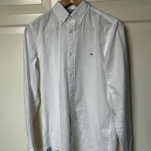 Vit skjorta från Tommy Hilfiger - Snygg vit skjorta från Tommy Hilfiger i slim fit med klassisk krage och knappar. Perfekt för en stilren look. Den har en liten broderad logotyp på bröstet. Är lite gul i armhålorna men inget som syns vid användning. Fraktar gärna, Finns och hämta i Bollebygd.                                                                                                      Hör av er vid frågor!