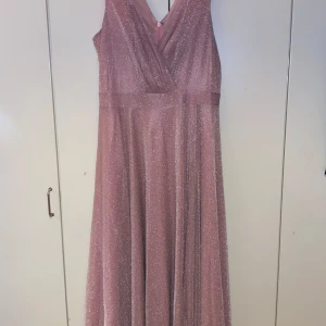 Rosa glittrig långklänning - Elegant rosa långklänning med glittrigt tyg och omlott-design. Klänningen är ärmlös och har en diskret dragkedja i ryggen. Och även en slits på vänster sida. Perfekt för festliga tillfällen. Använd 1 gång. Inköpt för 1299kr.