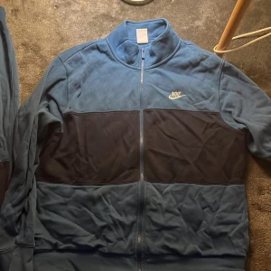 Blå träningsset från Nike - Säljer ett snyggt blått träningsset från Nike med dragkedja på tröjan och elastisk midja på byxorna. Tröjan har en mörkare blå panel över bröstet och Nike-logga på bröstet och benet. Perfekt för en sportig look!   Köpte för 999kr