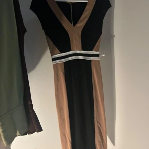 Svart och beige klänning med v-ringning - Elegant svart och beige klänning med djup v-ringning och kontrasterande paneler. Klänningen har en figurnära passform och ett stilrent band i midjan som framhäver silhuetten.