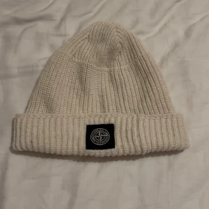 Vit stickad mössa från Stone Island - Säljer en snygg vit stickad mössa från Stone Island. Den har en ribbad design och en svart broderad logga framtill. Perfekt för att hålla sig varm under kyliga dagar. Passar både till vardags och mer avslappnade tillfällen.