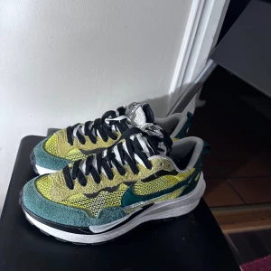 Nike sacai - Säljer ett par unika Nike sneakers i grönt och gult med svart snörning. Skorna har en cool, flerdimensionell sula och är perfekta för både vardag och träning. De är i bra skick och redo för nya äventyr. Passar perfekt för dig som vill sticka ut med stil!