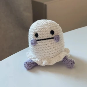 Litet spöke - En söt, handgjord virkad figur i vitt med lila detaljer. Den har en rund form med små lila fötter och kinder, samt en enkel svart mun och ögon. Perfekt som en liten dekoration eller present.