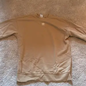 Säljer en snygg beige sweatshirt från Adidas. Den har en klassisk design med rund hals och långa ärmar. Perfekt för en avslappnad stil eller som lager under jackan. Adidas-loggan är diskret placerad på bröstet. Passar både till vardags och träning!