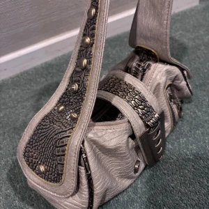 Beige axelväska med krokodilmönster - Säljer en cool beige axelväska med krokodilmönstrade detaljer och nitar. Väskan har flera dragkedjor och en unik design med metallspännen. Perfekt för att ge din outfit en unik touch! Leopardmönster inuti!!