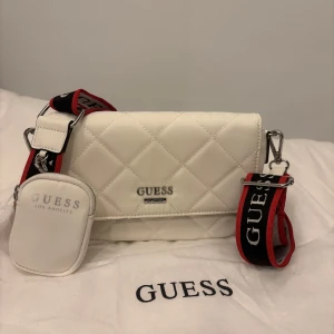 Vit quiltad axelväska från Guess - Säljer en superfin vit quiltad axelväska från Guess. Den är helt oanvänd! Den har en justerbar axelrem med Guess-logga i rött och svart. Väskan är i syntetmaterial och har en praktisk dragkedja inuti. Perfekt för både vardag och fest!