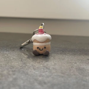 Nyckelring med cupcake-figur - Söt nyckelring med en cupcake-figur som har ett glatt ansikte. Baserat på jellycat gosedjuren! Cupcaken har vit glasyr, röda prickar och ett randigt ljus på toppen. Perfekt som en rolig accessoar till väskan eller nycklarna. Den är cirka 4 cm hög och har en praktisk karbinhake för enkel fastsättning.