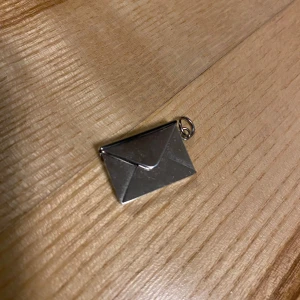 Silverfärgat kuvertberlock - Säljer en unik och stilren berlock i form av ett litet kuvert. Den är silverfärgad och har en liten ögla för att fästa på ett armband eller halsband. Perfekt för att ge en personlig touch till dina smycken. Berlocken är ca 2 cm lång och har en öppningsbar funktion. Passar perfekt som en liten hemlig förvaring eller bara som en söt detalj.