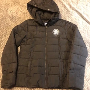 Svart pufferjacka från Svea - Säljer en svart pufferjacka från Svea i storlek L. Jackan har en dragkedja framtill och en broderad logga på bröstet. Perfekt för kyliga dagar med sin varma och bekväma design. Huvan ger extra skydd mot väder och vind. Passar både till vardags och lite mer avslappnade tillfällen. Det saknas ett bälte till midjan och päls på luvan, men inget som påverkar funktionen