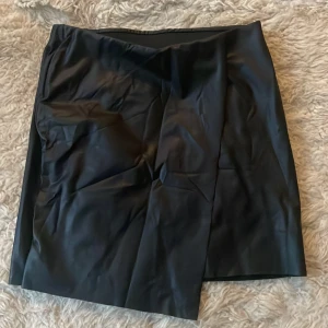 Svart kjol från Gina Tricot ny - Snygg svart kjol i skinnimitation från Gina Tricot. Perfekt för en kväll ute eller en stilren vardagslook. Kjolen har en enkel design med en liten slits framtill för extra stilpoäng. Passar bra till både höst och vintergarderoben.