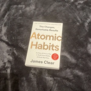 Atomic Habits - Säljer boken 'Atomic Habits' av James Clear. Den är en internationell bästsäljare som handlar om hur små förändringar kan leda till stora resultat. Perfekt för dig som vill bygga goda vanor och bryta dåliga. Boken har en vit framsida med titeln i stora orange bokstäver.