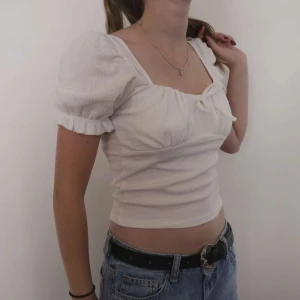 Vit kortärmad topp med puffärmar - Säljer en superfin vit topp med puffiga kortärmar och en söt knytdetalj framtill. Toppen är croppad och har en fyrkantig urringning som ger en romantisk känsla. Perfekt för sommardagar eller en mysig vårkväll. Passar bra till jeans eller kjol!