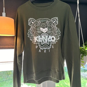 Grön sweatshirt från Kenzo - Säljer en snygg grön sweatshirt från Kenzo med det ikoniska tigermotivet. Tröjan är i storlek M och har långa ärmar. Perfekt för en casual look. Den är i bra skick, dock en fläck på bakre arm. Se bild.