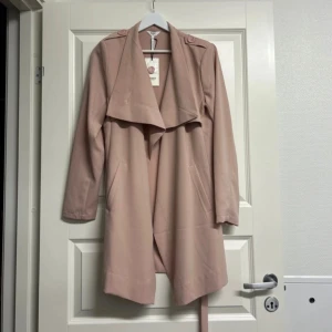 Beige trenchcoat med bälte - Säljer en snygg beige trenchcoat med bälte. Den har långa ärmar och knappar på axlarna för en stilren look. Perfekt för vår och höst!