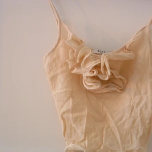 Beige linne från NA-KD - Säljer ett supersnyggt beige linne från NA-KD. Det är ärmlöst med smala axelband och har en fin volangdetalj framtill. Perfekt för sommardagar eller att styla med en kavaj för en chic look.