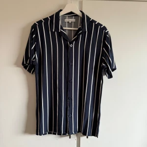 Svartvit randig skjorta från S.C.W. - Snygg svart skjorta med vita ränder från S.C.W. i storlek S. Skjortan har korta ärmar och knappar framtill, perfekt för en stilren look. Tillverkad i ett lätt och bekvämt material. ser lite konstig ut där bak där lappen sitter men det märks knappt på avstånd. Mycket bra skick annars. Aldrig använd. 