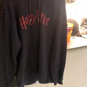 Mörkblå lång oversized Harry Potter hoodie - Säljer en svart hoodie med Harry Potter-tryck i rött. Den har en klassisk design med huva och snören. Perfekt för alla Potterheads som vill ha en bekväm och stilren tröja.