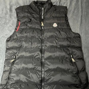 Svart dunväst från Moncler - Snygg svart dunväst från Moncler med dragkedja och logotyp på bröstet. Västen har en röd insida och praktiska fickor med dragkedja. Perfekt för kyligare dagar.