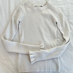 Vit ribbad tröja - Säljer en stilren vit ribbad tröja med långa ärmar och volangdetaljer vid ärmsluten. 