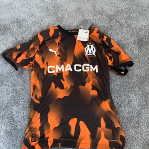 Orange och svart fotbollströja från Puma - Snygg fotbollströja från Puma i orange och svart med Marseille-logga och sponsortryck. Tröjan har korta ärmar och ett unikt mönster. Perfekt för fotbollsfans som vill sticka ut på planen.