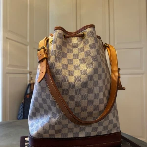 Rutig axelväska från Louis Vuitton - Snygg rutig axelväska modell Noé från Louis Vuitton i beige och grått.  Väskan har en justerbar rem i brunt skinn och detaljer i guld. Perfekt för att ge din outfit en lyxig touch. Kvitto finns! 
