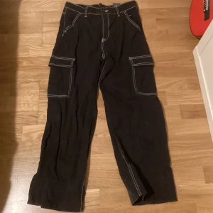 Svarta cargobyxor från H&M Divided - Svarta cargobyxor med vita sömmar från H&M Divided. Byxorna har flera praktiska fickor. Den har dock hål på högra byxbenet men annars är det inget fel på dem.