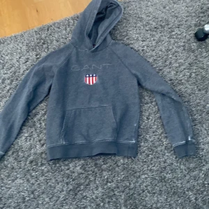 Grå hoodie från Gant - Säljer en snygg grå hoodie från Gant med en broderad logga på bröstet. Tröjan har en klassisk känguruficka och en bekväm huva. Perfekt för en avslappnad stil.