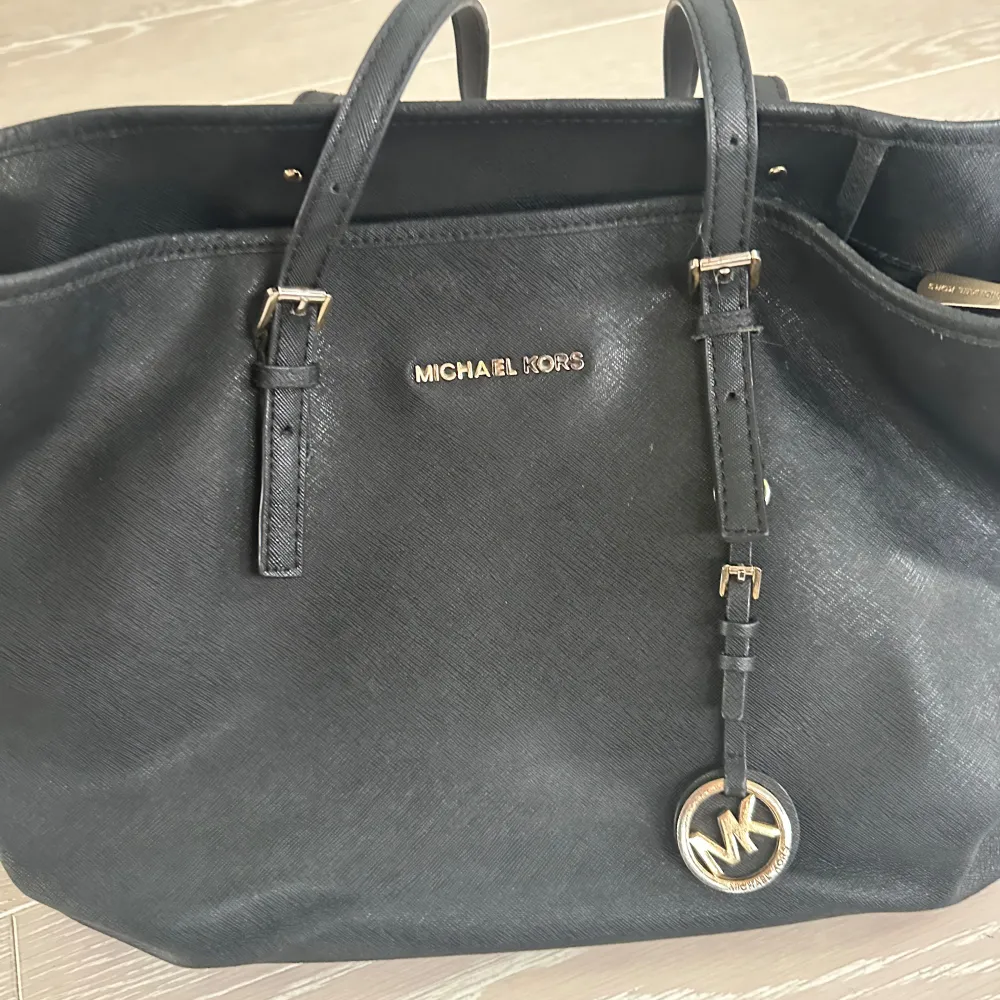Snygg svart axelväska från Michael Kors med guldfärgade detaljer och logotyp. Väskan har dubbla handtag och en dekorativ MK-berlock. Perfekt för att bära med sig allt man behöver med stil. Den är mycket väl använd! Se bilder för skick. Självklart äkta!. Laukut & Käsilaukut.