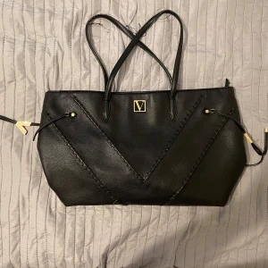 Svart axelväska med dragkedja - Snygg svart axelväska i skinn med elegant V-logga framtill. Väskan har dekorativa sömmar och guldfärgade detaljer. Insidan är randig och har en praktisk dragkedja för säker förvaring. Perfekt för att bära dina dagliga nödvändigheter med stil.