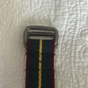 Randigt bälte från Polo - Säljer ett randigt bälte från Polo med en kombination av färgerna röd, grön och gul. Bältet har en metallspänne med Polo-logotypen ingraverad. Perfekt för att ge en färgklick till din outfit!