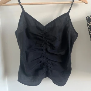 Svart ärmlös topp med rynkdetalj - Snygg svart ärmlös topp med smala axelband och rynkdetalj framtill. Perfekt för en stilren look. Oanvänd från H&M. perfekt till fest