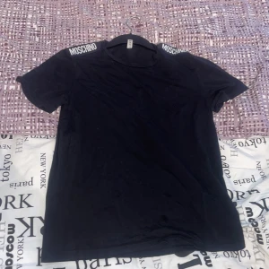 Svart t-shirt från Moschino - Snygg svart t-shirt från Moschino med logotyp på axlarna. Perfekt för en stilren look. Kortärmad och bekväm passform.