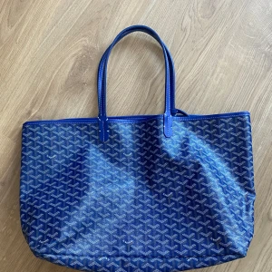 Blå mönstrad shoppingväska - Snygg blå shoppingväska med ett klassiskt mönster. Väskan har två handtag och en matchande liten plånbok med knappstängning. Insidan är ljus och rymlig, perfekt för att bära med sig allt man behöver. Perfekt för en dag på stan eller en shoppingrunda.