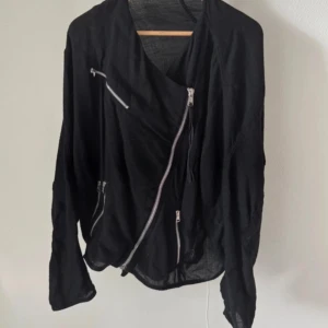 Svart jacka från Acne - Snygg svart jacka från Acne med asymmetrisk design och flera dragkedjor. Jackan har en cool och modern look, perfekt för att ge din outfit en edgy touch. Den är långärmad och tillverkad i ett lätt material.