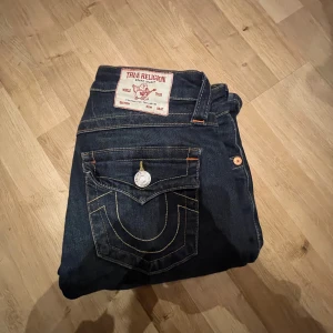 Mörkblå jeans från True Religion - Snygga mörkblå jeans från True Religion med klassisk femficksdesign och kontrastsömmar. Jeansen har en ikonisk bakficka med broderad detalj och en knappstängning fram. Perfekta för en stilren look.
