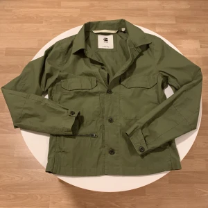 Grön jacka från G-Star RAW - Snygg grön jacka från G-Star RAW med knappar framtill och två bröstfickor. Perfekt för en stilren look. Jackan har långa ärmar och en klassisk krage.