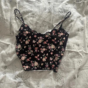 Blommig svart linnetopp - Söt svart linnetopp med blommigt mönster i rosa och grönt. Toppen har smala axelband och en lätt vågig kant upptill. Perfekt för varma dagar eller som ett lager under en kofta.
