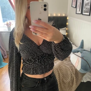 Svart och vit prickig blus - Säljer en snygg svart blus med vita prickar. Blusen har långa ärmar och en v-ringad design med knytning i midjan. 