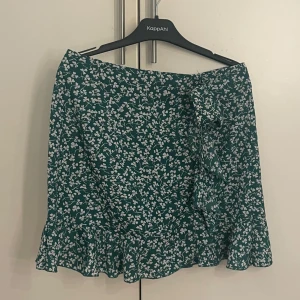 Grön blommig kjol - Söt grön kjol med vita blommor. Kjolen har en volangdetalj och knytning på sidan, vilket ger en fin omlott-effekt. Perfekt för sommarens varma dagar! Storlek M men har en liten passform.
