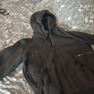 Svart fleecejacka från Sail Racing - Säljer en svart fleecejacka från Sail Racing med dragkedja och huva. Jackan har en stilren design med Sail Racing-logga på bröstet och är perfekt för kyligare dagar. Den är mjuk och bekväm att bära.