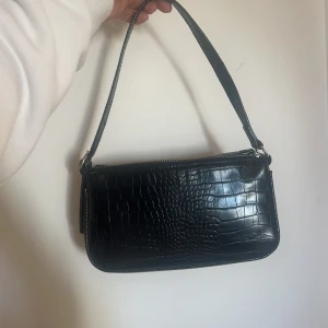 Svart axelväska med krokodilmönster - Snygg svart axelväska med krokodilmönster och justerbar rem. Väskan har en dragkedja upptill och en stilren design som passar perfekt för en trendig look. Perfekt för att bära dina nödvändigheter med stil.