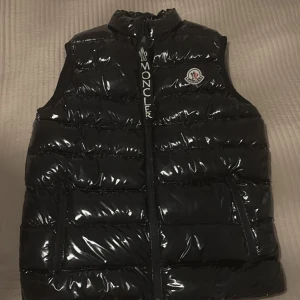 Svart dunväst från Moncler - Snygg svart dunväst från Moncler med glansig yta och dragkedja framtill. Västen har en broderad logotyp på bröstet och praktiska fickor. Perfekt för kyligare dagar när du vill hålla stilen.