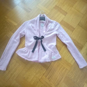 Rosa kofta från Odd Molly - Säljer en söt rosa kofta från Odd Molly med volangdetaljer och en svart rosett framtill. Koftan har långa ärmar och en mjuk, bekväm passform. Perfekt för att lägga till en feminin touch till din outfit. Storlek 1 passar xxs xs och s skulle jag säga  🌸