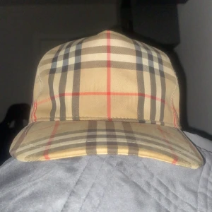 Rutig keps från Burberry - Snygg rutig keps från Burberry i klassiskt beige, svart och rött mönster. Kepsen har en broderad etikett med texten 'Burberry London England'. Perfekt för en stilren look.