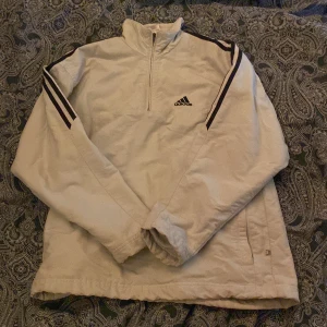 Vit jacka med dragkedja från Adidas - Säljer en vit jacka från Adidas med dragkedja och svarta ränder på axlarna. Jackan har en hög krage och är tillverkad i polyester. Perfekt för en sportig look!