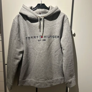 Grå hoodie från Tommy Hilfiger - Säljer en stilren grå hoodie från Tommy Hilfiger med broderad logga på bröstet. Hoodien har en mjuk insida och huva med dragsko. 