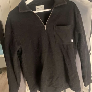 Svart tröja med dragkedja från Woodbird - Säljer en stilren svart tröja från Woodbird med dragkedja och bröstficka. Perfekt för kyligare dagar med sin långärmade design. Tröjan har en enkel och klassisk look.