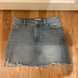 Blå jeanskjol från mango  - Snygg blå jeanskjol från mango med lite slitningar.  Kjolen har en kort längd och en knapp- och dragkedjestängning framtill. Kjolen är i så gott som mycket bra skick och den är använd ett fåtal gånger. Storlek 152. 