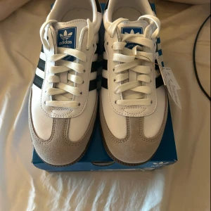 Adidas Samba OG - Säljer ett par Adidas Samba OG sneakers i vitt och svart lite beige aktig färg med snygga detaljer i mocka, brun sula, i storlek 39 1/3 tag och allting är kvar helt nya aldrig använda, köpta för 1449kr säljer då de är fel storlek för mig.